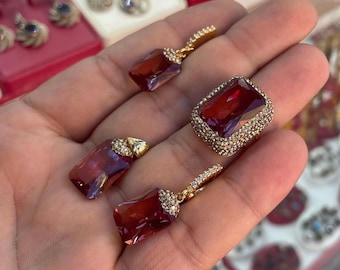 Puede incluir: Una colección de joyas doradas con piedras preciosas rectangulares de color rojo intenso. El conjunto incluye un anillo y dos pares de pendientes, todos con un halo de pequeñas piedras brillantes. Los pendientes tienen cierre de palanca.