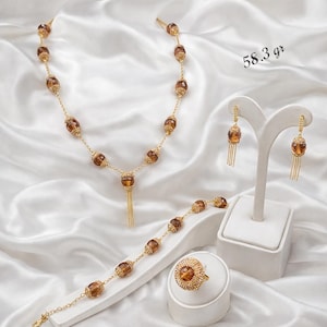 Puede incluir: Un conjunto de joyas dorado con collar, pulsera, pendientes y anillo. El conjunto incluye cuentas ovaladas de color ámbar y delicados eslabones de cadena. Los pendientes tienen borlas colgantes. El anillo tiene una gran piedra de color ámbar. El texto dice "58.3 gr".