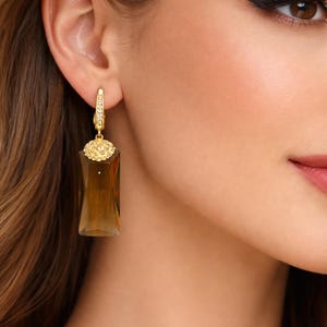 Puede incluir: Pendientes dorados con un colgante rectangular de piedra color ámbar. Los pendientes presentan un pequeño elemento redondo de oro texturizado en la parte superior y un cierre de bisagra. El diseño es elegante y adecuado para ocasiones formales.