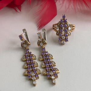 Puede incluir: Un conjunto de joyas doradas con piedras moradas y transparentes. El conjunto incluye un anillo y un par de pendientes colgantes.