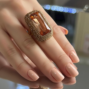 May include: A large, rectangular cocktail ring with a light orange gemstone and a gold-colored band. The ring is surrounded by smaller, gold-colored stones. The ring is on a hand with peach-colored nails.
