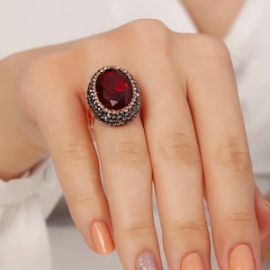 May include: A large, oval-shaped ruby-red gemstone ring. The ring features a dark red stone surrounded by a border of small, dark grey and gold-toned stones. The ring is set on a gold-coloured band. The hand has peach-coloured nail polish.