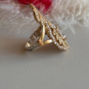 May include: A silver ring with a large, clear gemstone surrounded by a delicate, gold-toned, leaf-shaped design. The ring is marked with "925" on the band.