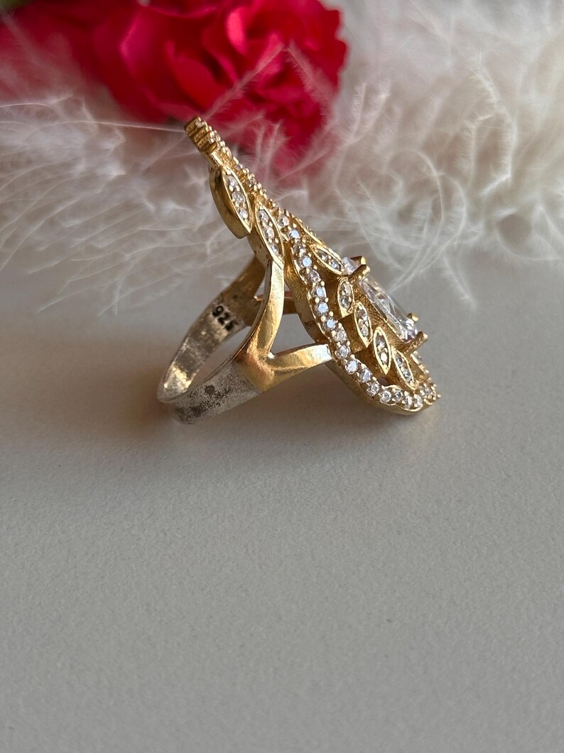 May include: A silver ring with a large, clear gemstone surrounded by a delicate, gold-toned, leaf-shaped design. The ring is marked with "925" on the band.