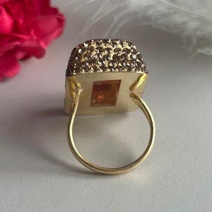 May include: A gold ring with a square-shaped stone set in a frame of small brown crystals. The ring is on a white surface.
