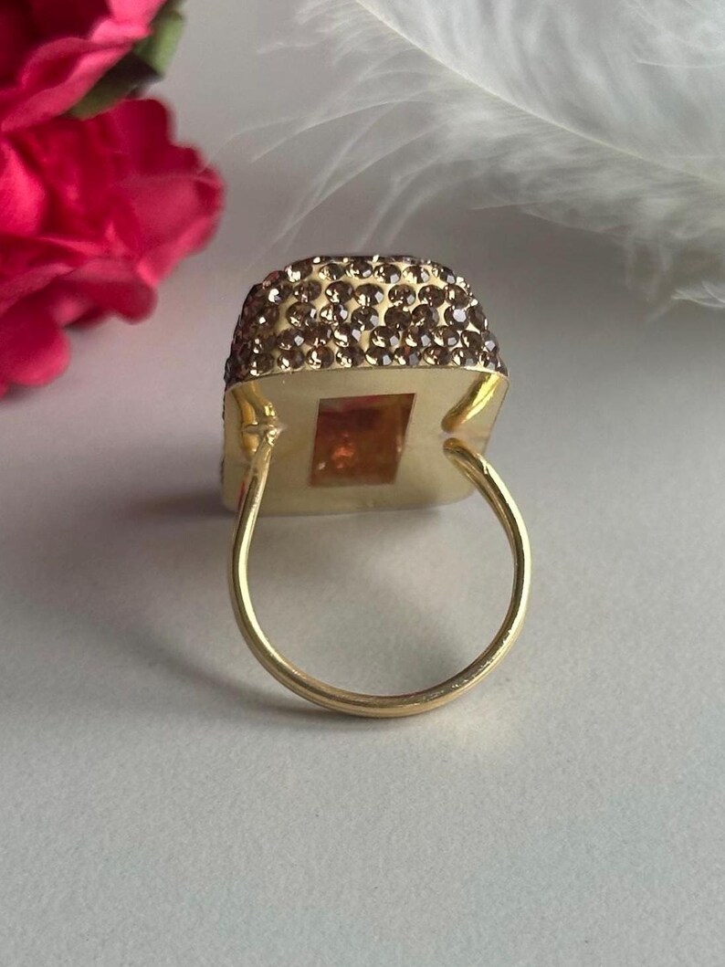 May include: A gold ring with a square-shaped stone set in a frame of small brown crystals. The ring is on a white surface.