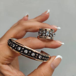 Puede incluir: Un brazalete tipo puño y un anillo a juego en tono oro rosa, ambos con una banda central negra texturizada incrustada con piedras claras de corte redondo. Las piezas de joyería tienen bordes lisos y pulidos en tono oro rosa.