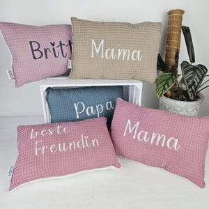 Könnte beinhalten: Fünf dekorative Kissen mit verschiedenen Stickereien. Die Kissen sind rosa, beige und blau. Der Text auf den Kissen lautet "Britt", "Mama", "Papa", "beste Freundin" und "Mama".