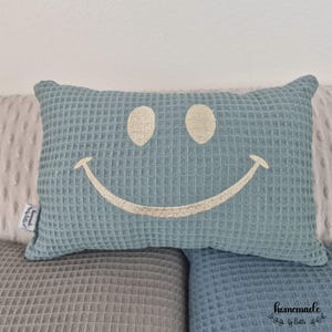 Puede incluir: Una almohada azul con textura de gofre y una cara sonriente bordada. La almohada rectangular descansa sobre una superficie texturizada gris y azul. La almohada tiene una etiqueta que dice "Homemade by Betti".