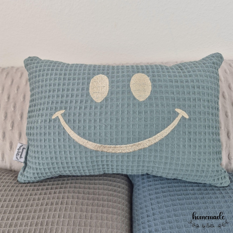 Puede incluir: Una almohada azul con textura de gofre y una cara sonriente bordada. La almohada rectangular descansa sobre una superficie texturizada gris y azul. La almohada tiene una etiqueta que dice "Homemade by Betti".