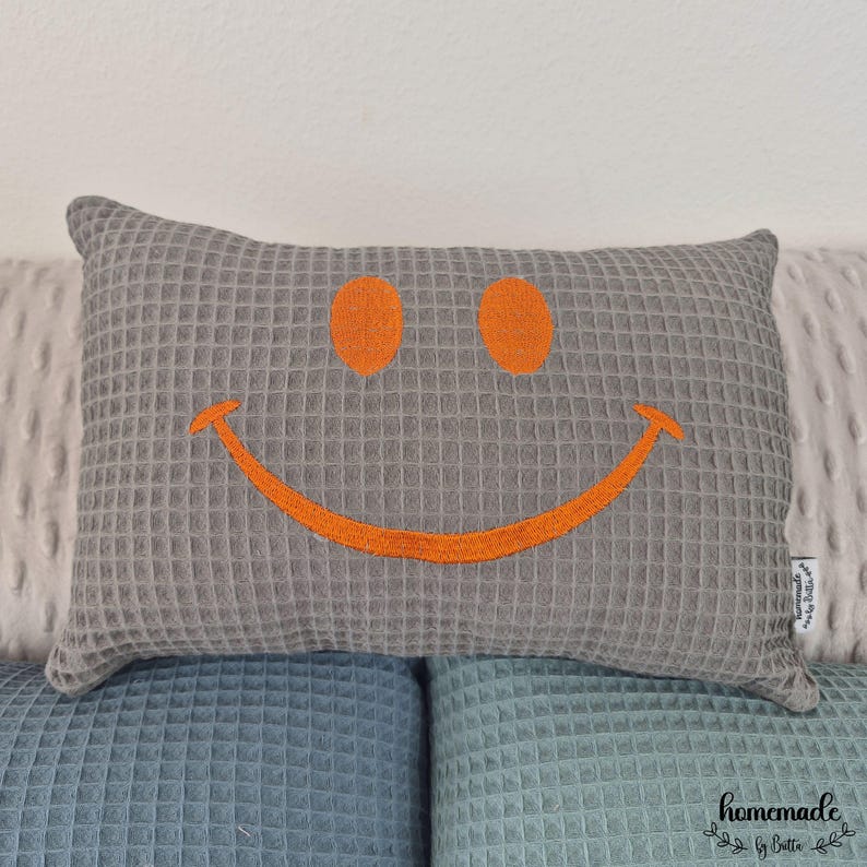Puede incluir: Coj&iacute;n rectangular gris con textura de gofre, con una cara sonriente naranja bordada. El coj&iacute;n est&aacute; colocado sobre una superficie texturizada azul y blanca. Un elemento decorativo para el hogar.