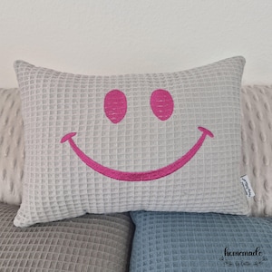 Puede incluir: Un coj&iacute;n rectangular gris claro con textura de gofre y una cara sonriente bordada en rosa brillante. El coj&iacute;n est&aacute; colocado encima de otros cojines en tonos grises y azules. Un art&iacute;culo decorativo.