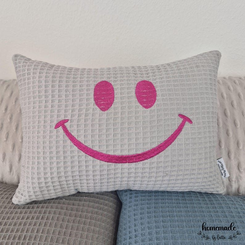 Puede incluir: Un coj&iacute;n rectangular gris claro con textura de gofre y una cara sonriente bordada en rosa brillante. El coj&iacute;n est&aacute; colocado encima de otros cojines en tonos grises y azules. Un art&iacute;culo decorativo.
