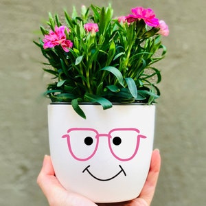 Op de afbeelding: Een witte keramische pot met een glimlachend gezicht en roze bril. De pot is gevuld met groene planten en roze bloemen.