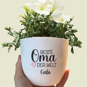 Könnte beinhalten: Ein weißer Keramik-Blumentopf mit dem Text "BESTE Oma DER WELT Gabi" in schwarzer Schrift, einem roten Herz und einer Pflanze mit weißen Blüten. Der Topf wird von einer Hand gehalten.