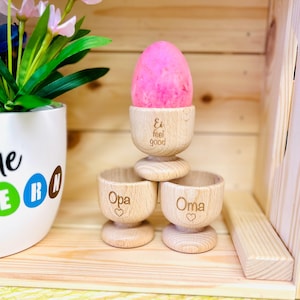 Op de afbeelding: Drie houten eierdopjes met de woorden "Ei feel good", "Opa" en "Oma" erop gegraveerd. Een roze ei zit in het bovenste dopje.