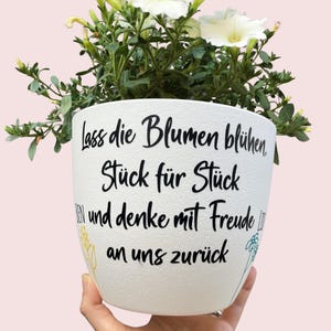 Może przedstawiać: Biała ceramiczna doniczka z czarnym napisem: "Lass die Blumen blühen, Stück für Stück, und denke mit Freude an uns zurück." Zawiera zielone liście i białe kwiaty. Doniczka jest trzymana przez osobę.
