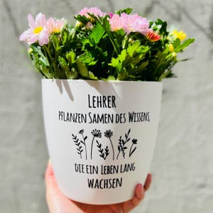 Könnte beinhalten: Ein weißer Keramik-Blumentopf gefüllt mit rosa und gelben Blumen und grünen Blättern. Der Topf hat einen schwarzen Text, der lautet: "LEHRER, PFLANZEN SAMEN DES WISSENS, DIE EIN LEBEN LANG WACHSEN" und ein Blumendesign.