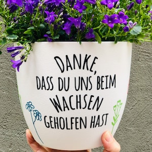Könnte beinhalten: Weißer Keramiktopf mit einer schwarzen Aufschrift "Danke, dass du uns beim wachsen geholfen hast". Der Topf ist mit grünen und blauen Blumenillustrationen verziert. Der Topf wird von einer Hand gehalten.