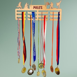 Może przedstawiać: Drewniany stojak na medale z wygrawerowanym imieniem "MILES". Stojak posiada wycięte figurki sportowców i mieści kilka medali z kolorowymi wstążkami. Medale są w kolorze złotym i mają różne wzory.