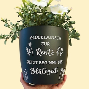 Könnte beinhalten: Ein dunkelgrüner Keramik-Blumentopf mit weißem Text, der lautet: "Glückwunsch zur Rente. Jetzt beginnt die Blütezeit." Der Topf ist mit grünen Pflanzen und weißen Blumen gefüllt. Der Topf wird von einer Person gehalten.