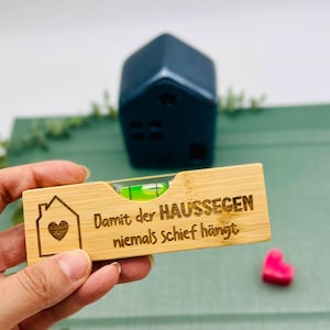 Op de afbeelding: Een houten waterpas met een bel, met een huisafbeelding met een hart en de Duitse tekst "Damit der HAUSSEGEN niemals schief hängt." Een donkerblauw huisvormig object staat op de achtergrond, samen met een klein roze hart.