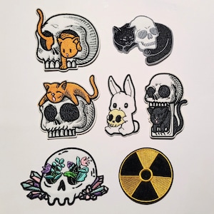 Peut inclure: Collection de patchs brodés avec des motifs gothiques et mignons. Les patchs incluent des crânes, des chats, un lapin et un symbole de radiation. Les patchs présentent un crâne avec un chat, un chat noir avec un crâne et un lapin tenant un crâne.