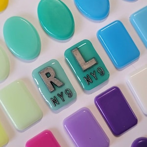 May include: A collection of smooth, glossy, rectangular and oval glass cabochons in various colours including teal, blue, purple, and white. Two teal rectangular pieces feature the silver-coloured letters R and L with the text NY9 printed below.