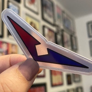 Puede incluir: Una pegatina reflectante plateada con un diseño de flecha roja, blanca y azul.