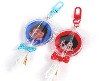 May include: Two lollipop-shaped keychains. One has a blue background and a blue clip, the other has a red background and a red clip. Both have cartoon character faces, bows, and gold-colored tassels.