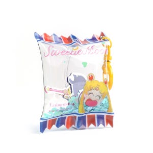 Peut inclure: Une pochette transparente en forme de bonbon avec un clip jaune. La pochette présente une image de dessin animé d'un personnage aux cheveux jaunes et le texte "Sweetie Moon" et "Eating makes me so happy". La pochette a des accents rouges, blancs et bleus.