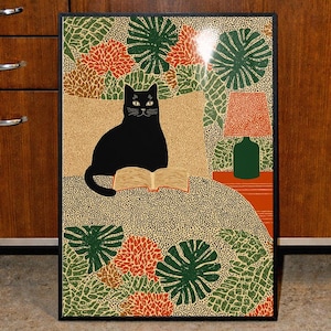 May include: A framed art print featuring a black cat reading a book, set against a patterned background with green leaves and orange flowers. A lamp and a green bottle are on a side table. The artwork has a black frame.