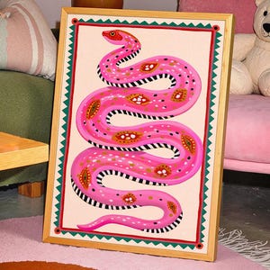 May include: A framed art print featuring a stylized pink snake with decorative patterns. The snake is set against a cream background, framed with a green and red border, and a natural wood frame. The artwork is displayed in a room with a pink rug and green sofa.