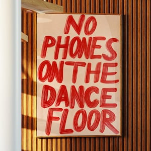 May include: A framed art print with the text "NO PHONES ON THE DANCE FLOOR" in red lettering. The artwork hangs on a vertical wood-paneled wall. A spiral staircase and a glimpse of a seating area are also visible.