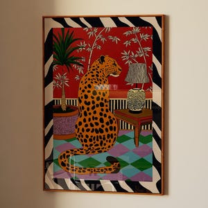 May include: A framed art print featuring a stylized leopard with orange spots, set against a red background with a black and white striped border. The artwork includes potted plants, a small table, and a patterned rug, creating a vibrant and decorative scene.