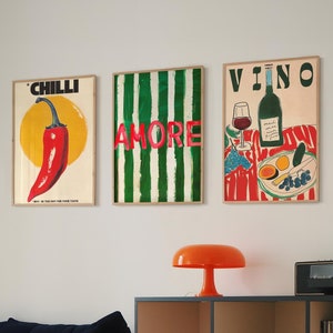 May include: Three framed prints with colourful designs. The first print has a red chilli pepper on a white background with the text "CHILLI". The second print has green and white stripes with the text "AMORE" in red. The third print has a red and white checkered background with a bottle of wine, a glass of wine, and fruit. The text "VINO" is at the top of the print.