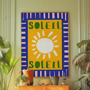 Puede incluir: Una impresión enmarcada en amarillo, azul y rosa con la palabra "SOLEIL" escrita dos veces en letras verdes. La impresión presenta un sol blanco con rayos amarillos sobre un fondo amarillo.