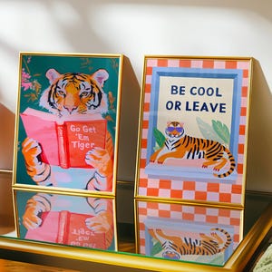 May include: Two rectangular framed art prints with gold borders. The left print shows a tiger reading a red book titled Go Get 'Em Tiger. The right print features a tiger wearing sunglasses with the text BE COOL OR LEAVE on a checkered background.