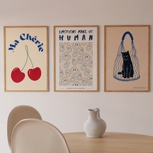 May include: Three framed prints with a minimalist design. The first print features a red cherry with the text "Ma Cherie" in blue. The second print has a pattern of blue smiley faces on a beige background with the text "Emotions Make Us Human" in blue. The third print features a black cat in a blue net bag on a beige background with the text "Artevina Design" in blue.