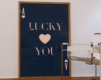 May include: A framed print with a navy blue background and a white heart with the text "LUCKY YOU" in white. The ace of hearts is in the top left and bottom right corners of the print.