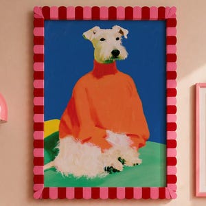 May include: A framed artwork depicts a dog wearing an orange sweater against a blue backdrop. The frame is pink and red. Below, striped and solid pillows in pink and red rest on a striped bedspread. A pink wall lamp is visible.
