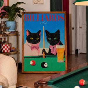 Funny cats playing pool poster featuring two black-faced cats wearing pink patterned bows behind a bright green billiards table. One tuxedo cat has a white body and yellow eyes, while the other is black with green eyes and holds an orange pool cue. Red and black billiard balls marked 8 and 7 sit on the felt beside a glass of golden beer. Large red BILLIARDS lettering, delicate Club script stand out against a vivid blue background. This quirky cat artwork suits game room, home bar, gallery wall.