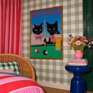 Funny cats playing pool poster featuring two black-faced cats wearing pink patterned bows behind a bright green billiards table. One tuxedo cat has a white body and yellow eyes, while the other is black with green eyes and holds an orange pool cue. Red and black billiard balls marked 8 and 7 sit on the felt beside a glass of golden beer. Large red BILLIARDS lettering, delicate Club script stand out against a vivid blue background. This quirky cat artwork suits game room, home bar, gallery wall.