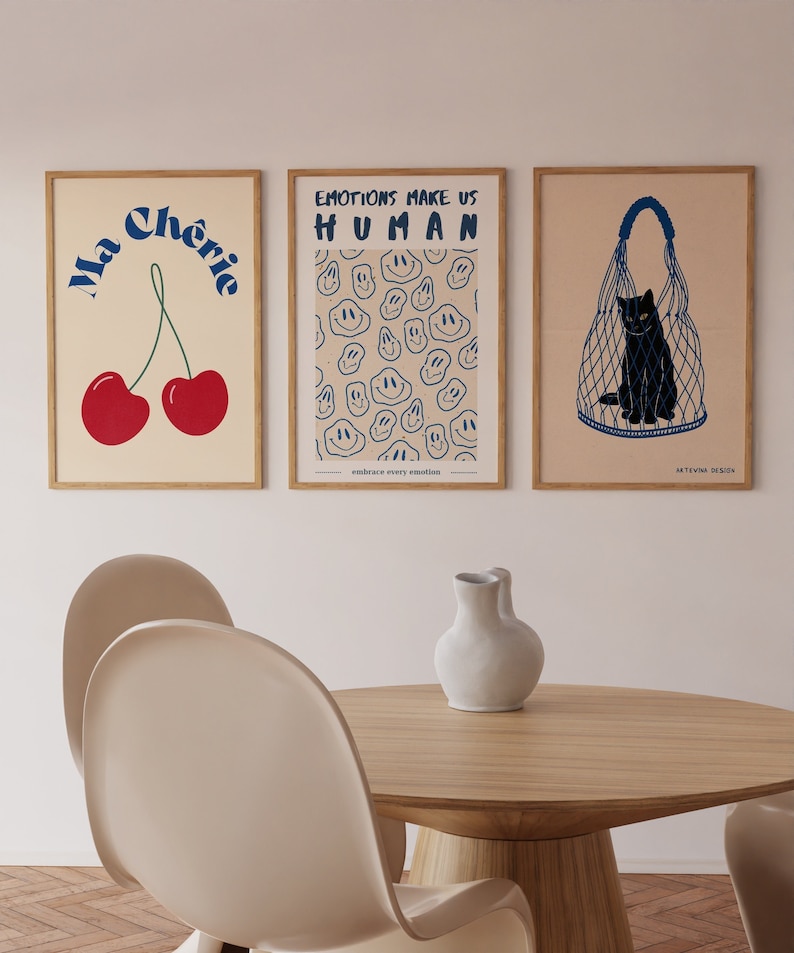 May include: Three framed prints with a minimalist design. The first print features a red cherry with the text "Ma Cherie" in blue. The second print has a pattern of blue smiley faces on a beige background with the text "Emotions Make Us Human" in blue. The third print features a black cat in a blue net bag on a beige background with the text "Artevina Design" in blue.