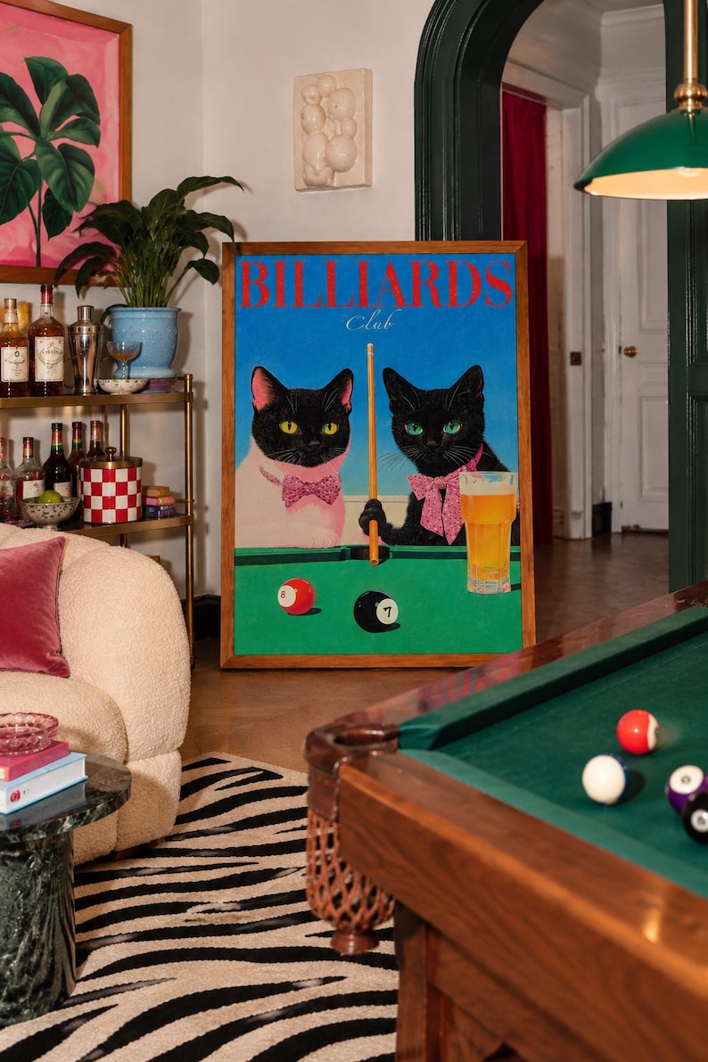 Funny cats playing pool poster featuring two black-faced cats wearing pink patterned bows behind a bright green billiards table. One tuxedo cat has a white body and yellow eyes, while the other is black with green eyes and holds an orange pool cue. Red and black billiard balls marked 8 and 7 sit on the felt beside a glass of golden beer. Large red BILLIARDS lettering, delicate Club script stand out against a vivid blue background. This quirky cat artwork suits game room, home bar, gallery wall.