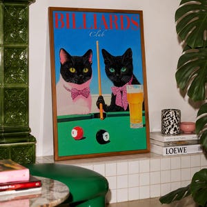 Funny cats playing pool poster featuring two black-faced cats wearing pink patterned bows behind a bright green billiards table. One tuxedo cat has a white body and yellow eyes, while the other is black with green eyes and holds an orange pool cue. Red and black billiard balls marked 8 and 7 sit on the felt beside a glass of golden beer. Large red BILLIARDS lettering, delicate Club script stand out against a vivid blue background. This quirky cat artwork suits game room, home bar, gallery wall.