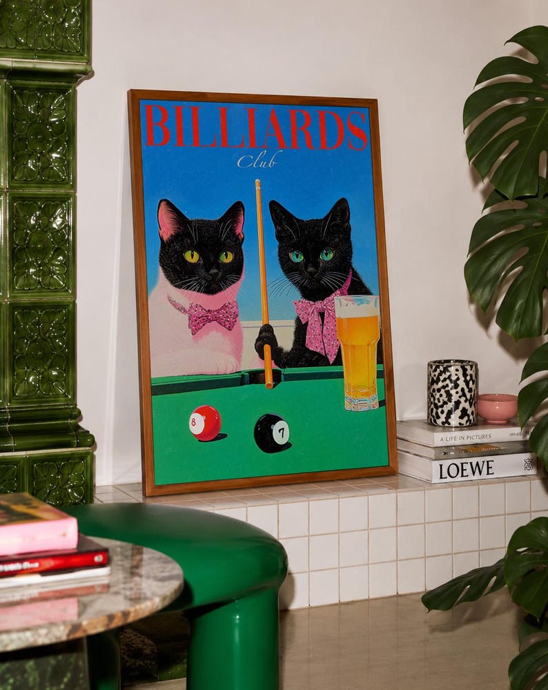 Funny cats playing pool poster featuring two black-faced cats wearing pink patterned bows behind a bright green billiards table. One tuxedo cat has a white body and yellow eyes, while the other is black with green eyes and holds an orange pool cue. Red and black billiard balls marked 8 and 7 sit on the felt beside a glass of golden beer. Large red BILLIARDS lettering, delicate Club script stand out against a vivid blue background. This quirky cat artwork suits game room, home bar, gallery wall.