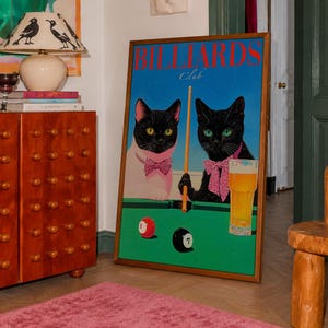 Funny cats playing pool poster featuring two black-faced cats wearing pink patterned bows behind a bright green billiards table. One tuxedo cat has a white body and yellow eyes, while the other is black with green eyes and holds an orange pool cue. Red and black billiard balls marked 8 and 7 sit on the felt beside a glass of golden beer. Large red BILLIARDS lettering, delicate Club script stand out against a vivid blue background. This quirky cat artwork suits game room, home bar, gallery wall.
