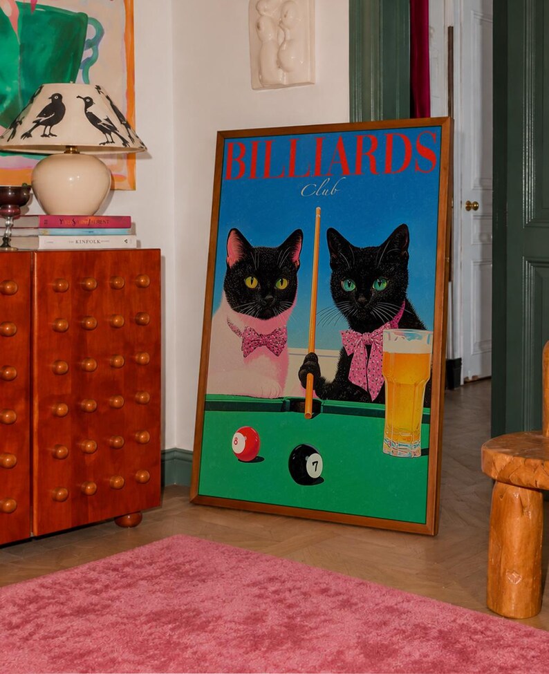 Funny cats playing pool poster featuring two black-faced cats wearing pink patterned bows behind a bright green billiards table. One tuxedo cat has a white body and yellow eyes, while the other is black with green eyes and holds an orange pool cue. Red and black billiard balls marked 8 and 7 sit on the felt beside a glass of golden beer. Large red BILLIARDS lettering, delicate Club script stand out against a vivid blue background. This quirky cat artwork suits game room, home bar, gallery wall.