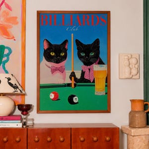 Funny cats playing pool poster featuring two black-faced cats wearing pink patterned bows behind a bright green billiards table. One tuxedo cat has a white body and yellow eyes, while the other is black with green eyes and holds an orange pool cue. Red and black billiard balls marked 8 and 7 sit on the felt beside a glass of golden beer. Large red BILLIARDS lettering, delicate Club script stand out against a vivid blue background. This quirky cat artwork suits game room, home bar, gallery wall.